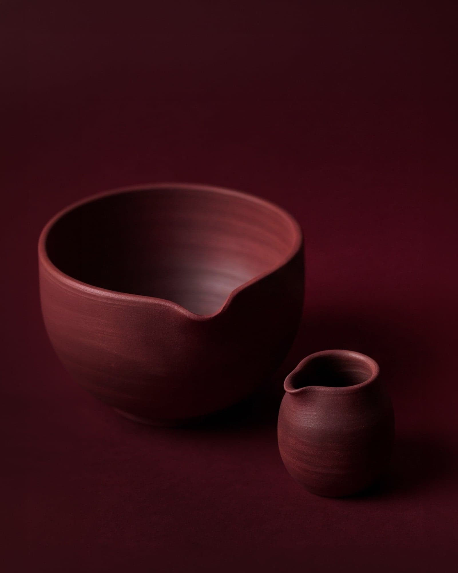 Two handmade burgundy ceramic pieces, a bowl and a small jug, on a deep red background